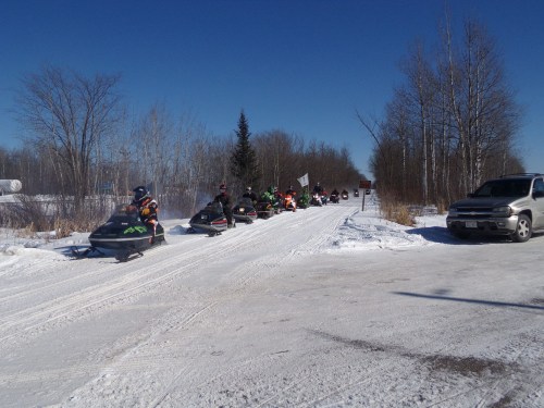 A line of snowmobiles on a snowy road, surrounded by trees under a clear blue sky. A parked vehicle is nearby.