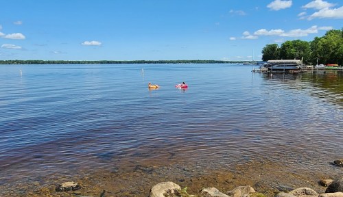 Two people float on colorful rafts in a calm lake, with a dock and trees in the background under a clear blue sky.