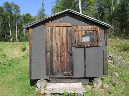 A small, rustic wooden cabin with a dark exterior, featuring a wooden door and a window, surrounded by greenery.