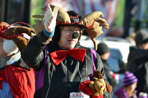 A woman in festive reindeer attire waves, holding a stuffed toy, surrounded by a cheerful crowd at an outdoor event.