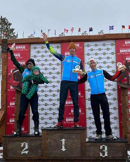 Three skiers celebrate on a podium, holding medals, with a child in a green outfit on one skier's arm. Flags in the background.