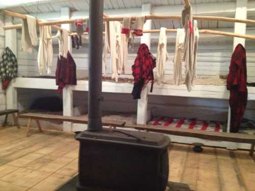 Interior of a rustic cabin with wooden bunks, hanging clothes, and a stove, creating a cozy, lived-in atmosphere.