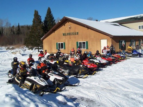 A group of snowmobilers in winter gear poses with their snowmobiles in front of a wooden building in a snowy landscape.
