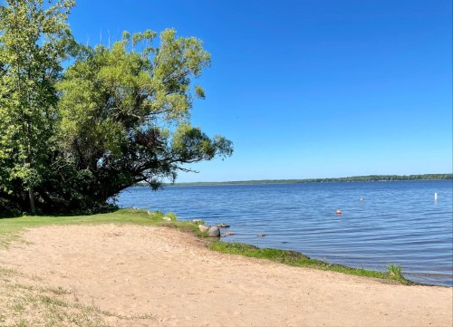 A sandy beach by a calm lake, surrounded by trees under a clear blue sky.