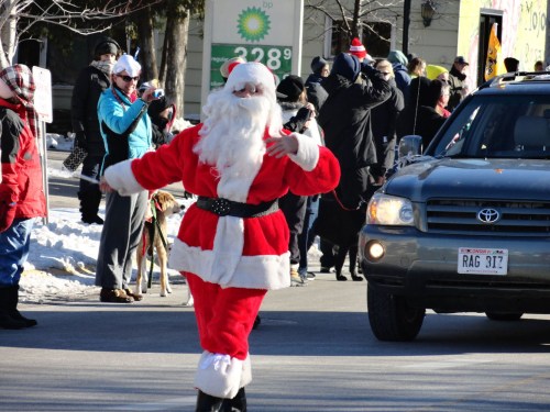 A person dressed as Santa Claus walks in a parade, with onlookers and a car in the background on a snowy street.