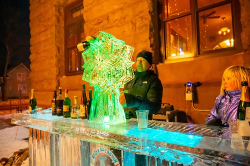 A bartender carves an illuminated ice sculpture at a winter event, with drinks displayed on an ice bar.
