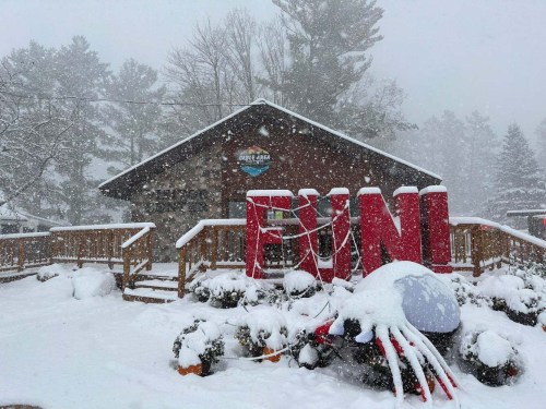 A snowy scene featuring a wooden building with a large red "FUN" sign in front, surrounded by snow-covered plants.