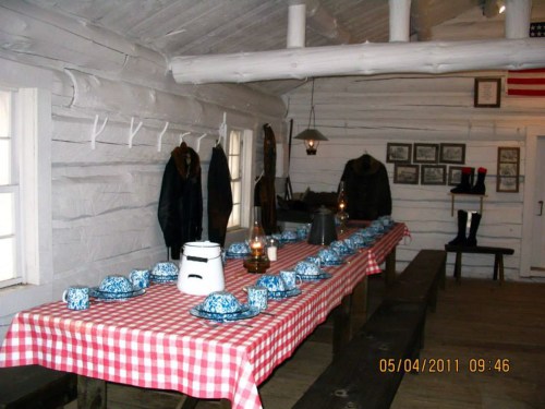 A rustic dining room with a checkered tablecloth, set with blue and white dishes, lanterns, and coats hanging on the wall.