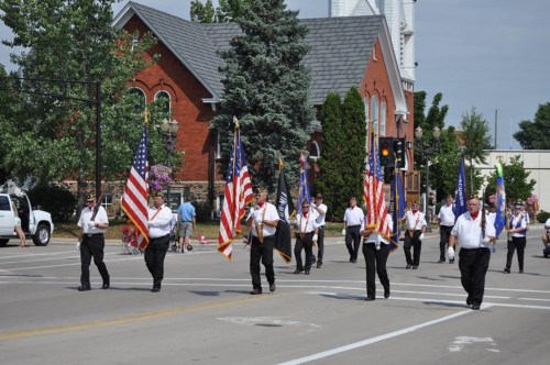 A parade featuring individuals in uniforms carrying flags, with a church and trees in the background.