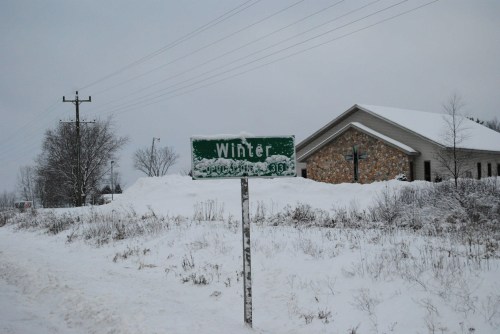 A snow-covered sign reading "Winter" next to a house, with a snowy landscape and overcast sky in the background.