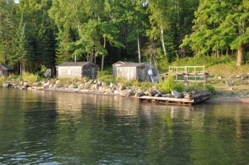 A serene lakeside scene featuring rustic cabins and lush greenery along the shore, reflecting in calm waters.