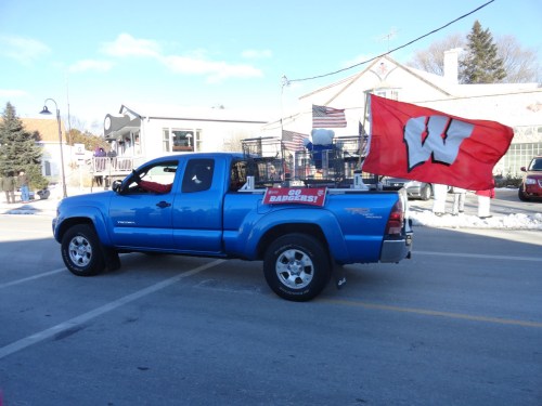 A blue pickup truck with a Wisconsin flag and "Go Badgers!" banner drives down a snowy street.