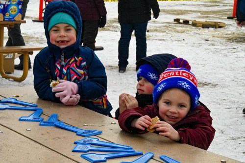 Two smiling children in winter clothing sit at a table, enjoying snacks, with blue game pieces scattered in front of them.