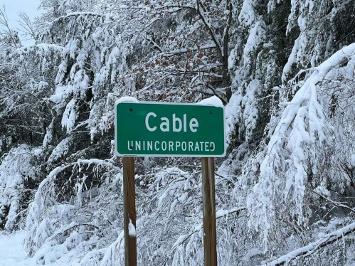 A green road sign reading "Cable UNINCORPORATED" surrounded by snow-covered trees.
