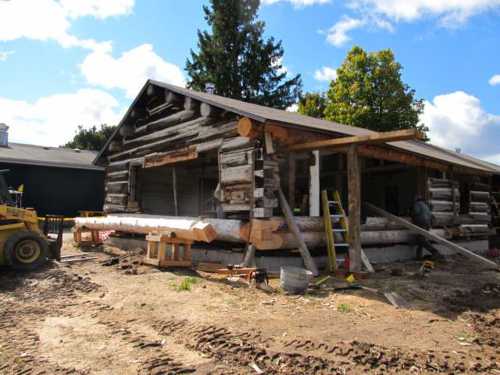 A log cabin under construction, with wooden beams and tools visible, surrounded by dirt and trees under a blue sky.