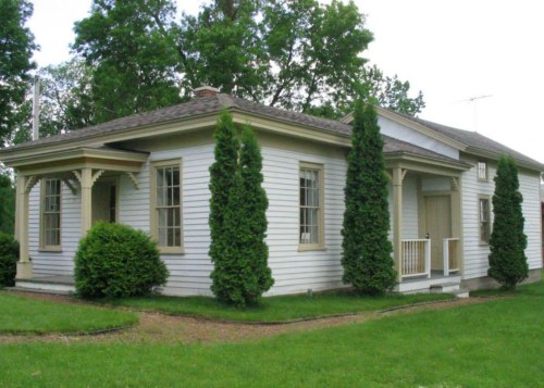A charming white house with green shrubs and tall trees, featuring a porch and multiple windows.