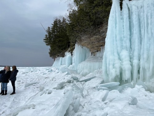 Two people stand on a snowy shore near icy cliffs and frozen formations, with a cloudy sky above.