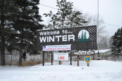 Sign welcoming visitors to Winter, featuring a tribute to astronaut Jeff Williams and surrounded by snow-covered trees.