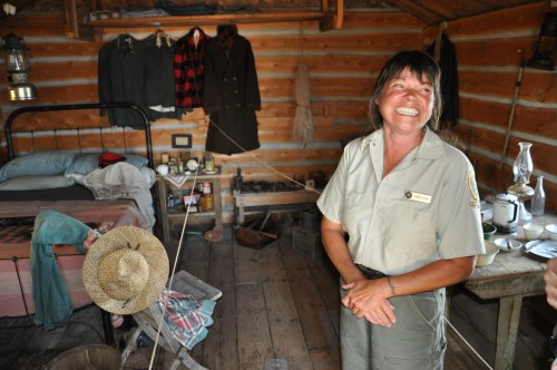 A smiling woman in a uniform stands in a rustic cabin with a bed, clothing on the wall, and various vintage items.