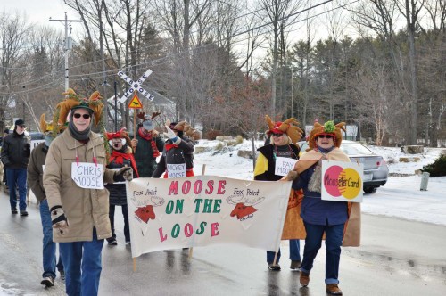 A festive parade with participants wearing moose antlers, holding a banner that reads "Moose on the Loose." Snowy setting.