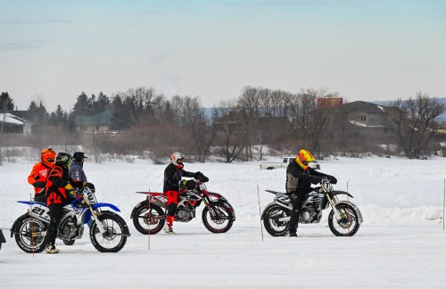 Four motocross riders prepare on snow-covered terrain, with trees and houses in the background under a clear sky.