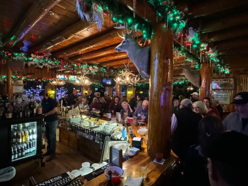 A lively bar scene with festive decorations, patrons enjoying drinks, and a mounted deer head on the wall.