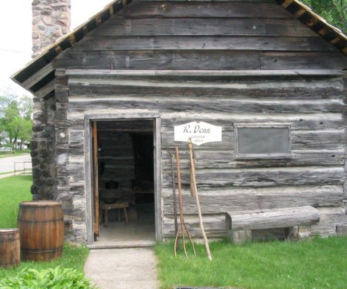 A rustic log cabin with a sign reading "R. Dunn" and wooden tools outside, surrounded by green grass and a barrel.