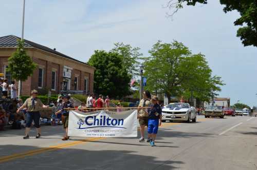 A parade scene with participants carrying a banner, surrounded by spectators and vehicles on a sunny day.