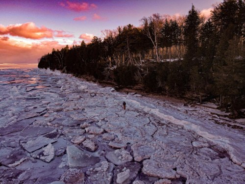 A person walks along a frozen shoreline with ice chunks and trees under a colorful sunset sky.