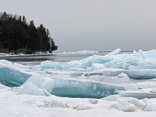 A winter landscape featuring broken ice formations on a frozen lake, with a forested shoreline in the background.