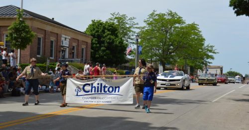 A parade scene with participants carrying a banner for the Chilton Chamber of Commerce, surrounded by spectators.