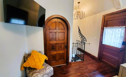 Cozy entryway featuring a wooden door, staircase, TV, and a chair with a yellow throw pillow. Warm lighting from a chandelier.