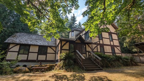 A charming, two-story house with a mix of wood and stone, surrounded by trees and greenery in a serene outdoor setting.