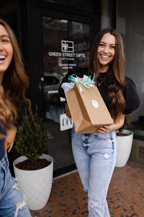 A smiling woman holds a gift bag outside a shop, with another woman laughing in the background.