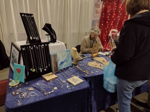 A vendor displays handmade jewelry at a market, while a customer browses and holds a blue shopping bag.