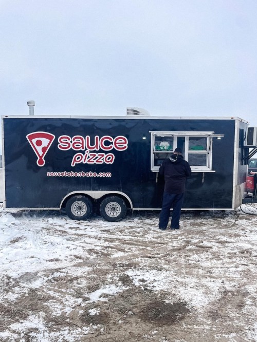 A person orders from a blue pizza food truck with a "sauce pizza" logo, set in a snowy outdoor location.