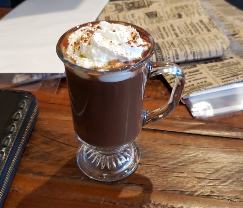A glass mug filled with hot chocolate topped with whipped cream and chocolate shavings, resting on a wooden table.