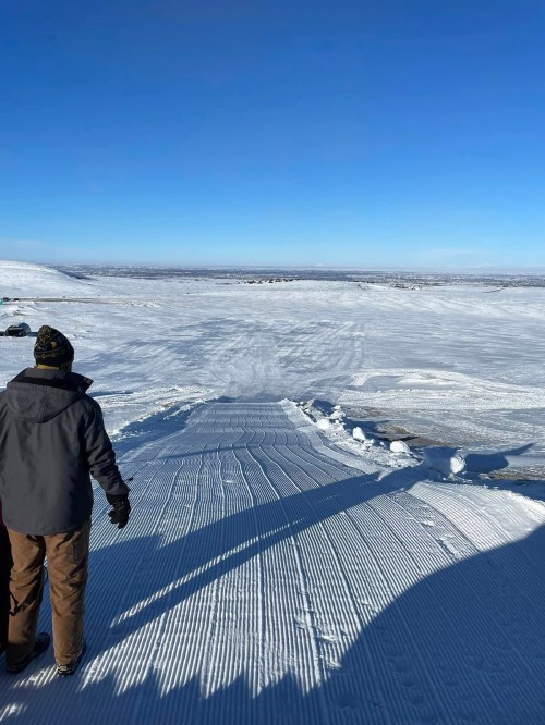 A person stands on a snowy landscape, looking out over a vast, clear blue sky and a horizon of untouched snow.