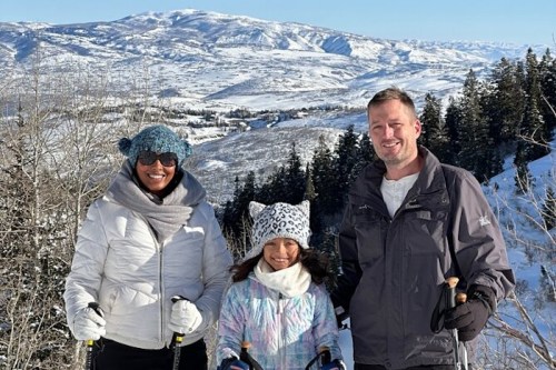 A family poses in winter gear on a snowy mountain, with trees and hills in the background.