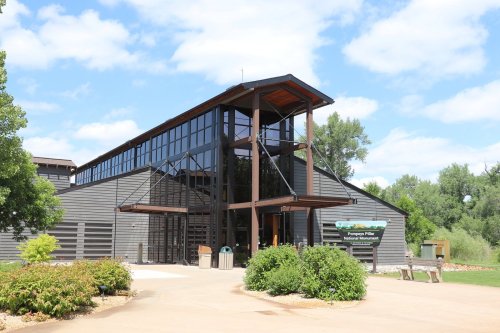 Modern building with large glass windows, surrounded by greenery and a pathway, under a blue sky with clouds.