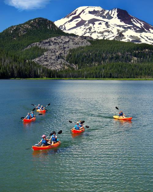 A group of kayakers paddles on a serene lake with a snow-capped mountain in the background.