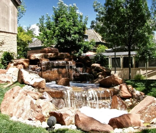 A serene waterfall cascades over rocks in a lush garden, surrounded by greenery and a clear blue sky.