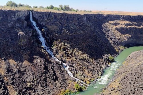 A waterfall cascades down rocky cliffs into a winding green river below, surrounded by dry land and sparse vegetation.