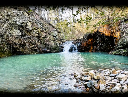 A serene scene of a turquoise pool with a small waterfall, surrounded by rocky terrain and lush greenery.