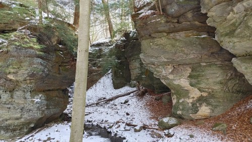 A serene winter landscape featuring rocky cliffs, snow-covered ground, and a small stream surrounded by trees.