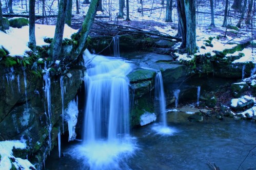 A serene winter scene featuring a waterfall cascading over rocks, surrounded by snow and icicles in a forested area.
