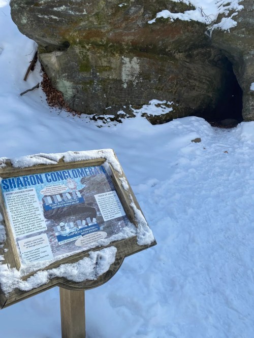 A snowy landscape featuring a sign about Sharon Conglomerate near a cave entrance in a rocky area.