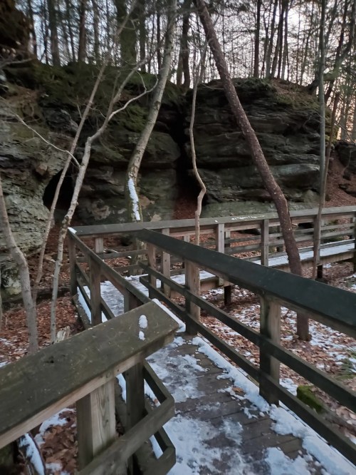A wooden walkway leads through a forest with rocky cliffs and snow-covered ground. Trees surround the path.