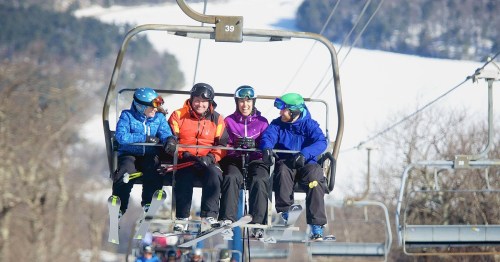 Four skiers on a chairlift, smiling and wearing colorful winter gear, with snowy slopes in the background.