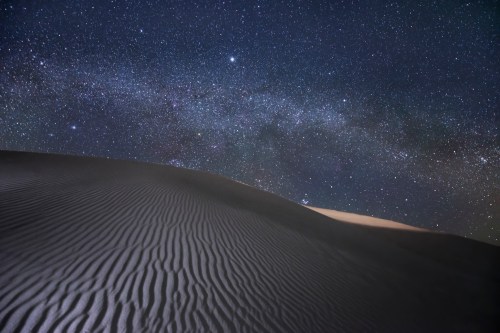 A serene desert landscape at night, featuring rippled sand dunes under a starry sky with the Milky Way visible.